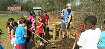 Students on a field trip to a farm are shown how to manually till earth by a volunteer.